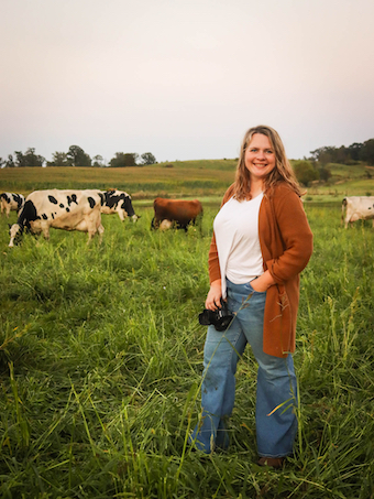 Brittany Olson standing in pasture at Berglane Farm, holding a camera, with dairy cows grazing behind her.
