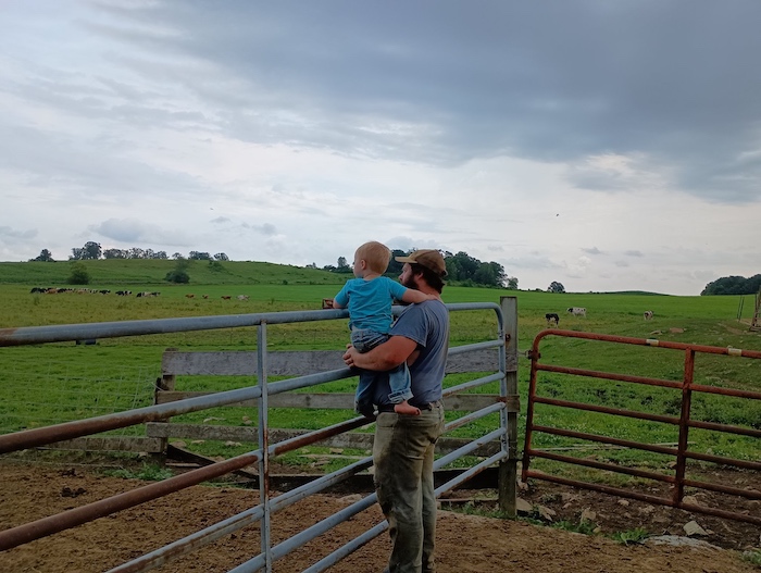 Sam Olson holding his young son at a farm gate, looking out over pasture at Berglane Farm in Wisconsin.