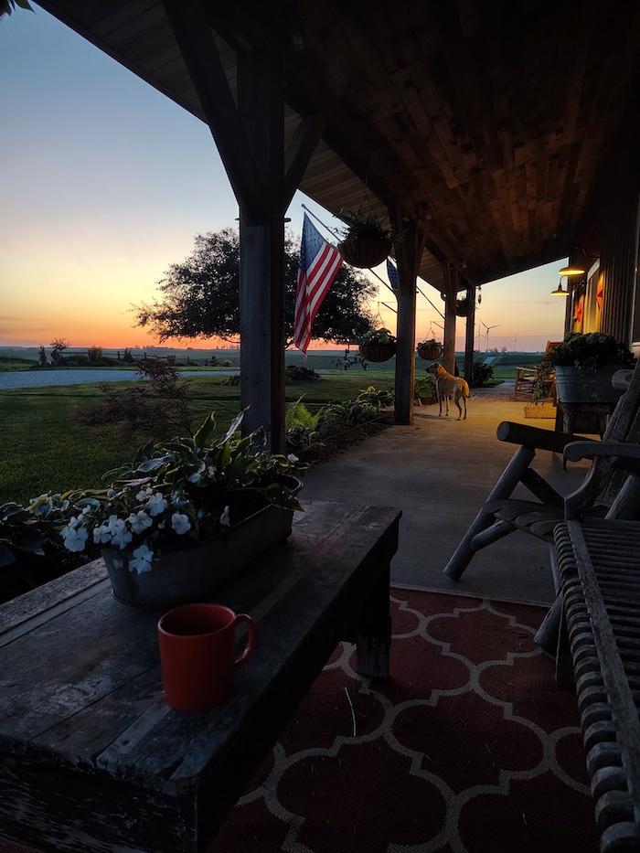 A sunrise view from the Yenter family porch, with a coffee cup on the table, an American flag, and the farm stretching out beyond.